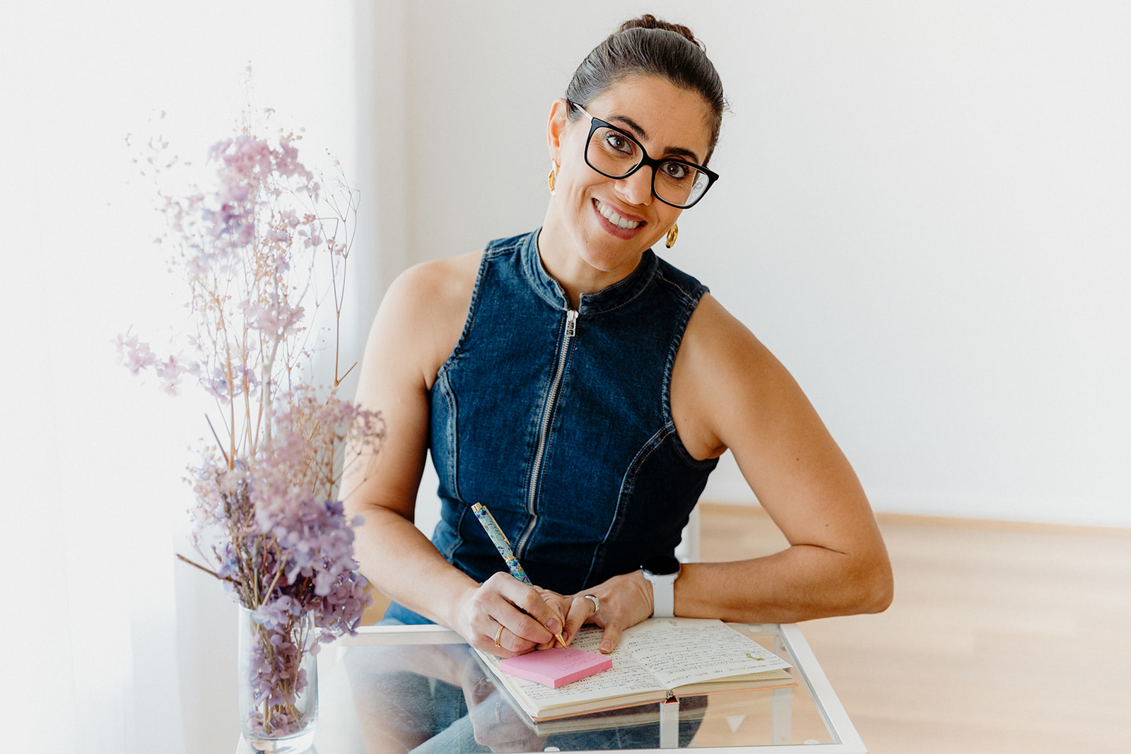 Sofía Silva Berenguer Sofía at a desk next to a window. On the desk there are some violet flowers. Sofía has her hands on a notebook and she is holding a pen on her right hand writing on pink sticky notes resting on top of the notebook. Sofía is wearing a denim sleeveless top zippered up to the neck.